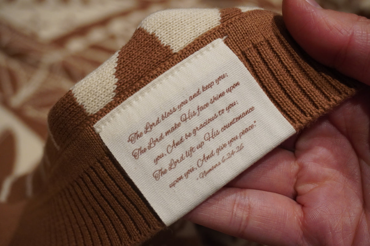 A close-up of a hand holding a brown and cream knit sweater cuff, showing a sewn-on cream fabric label with a printed Bible verse.