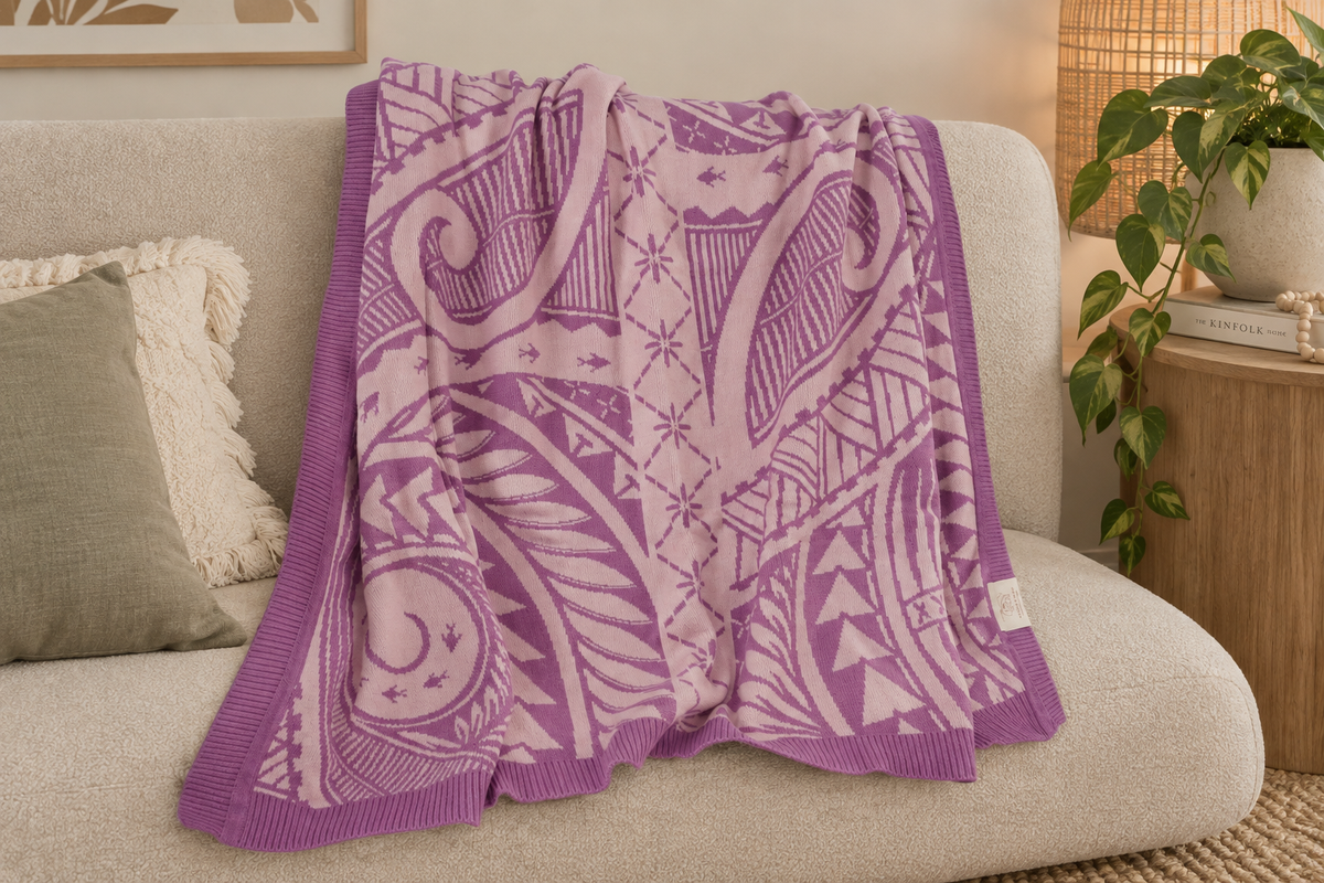 A purple and light pink knit throw blanket with pacific pattern, draped over a beige fabric sofa in a cozy living room setting.
