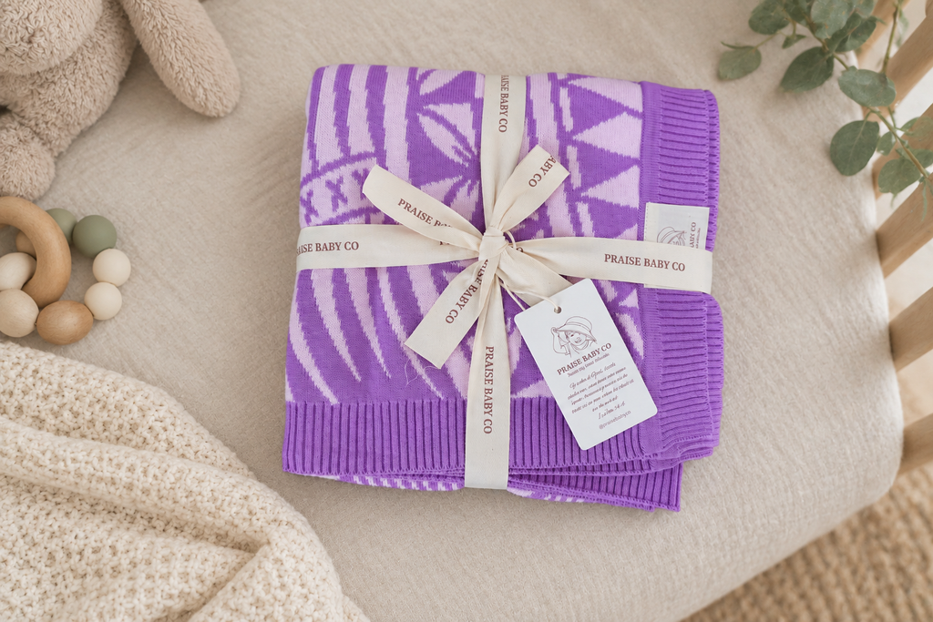 A folded purple and white patterned knit baby blanket tied with a cream ribbon and tag, styled on a beige fabric surface with soft nursery props.