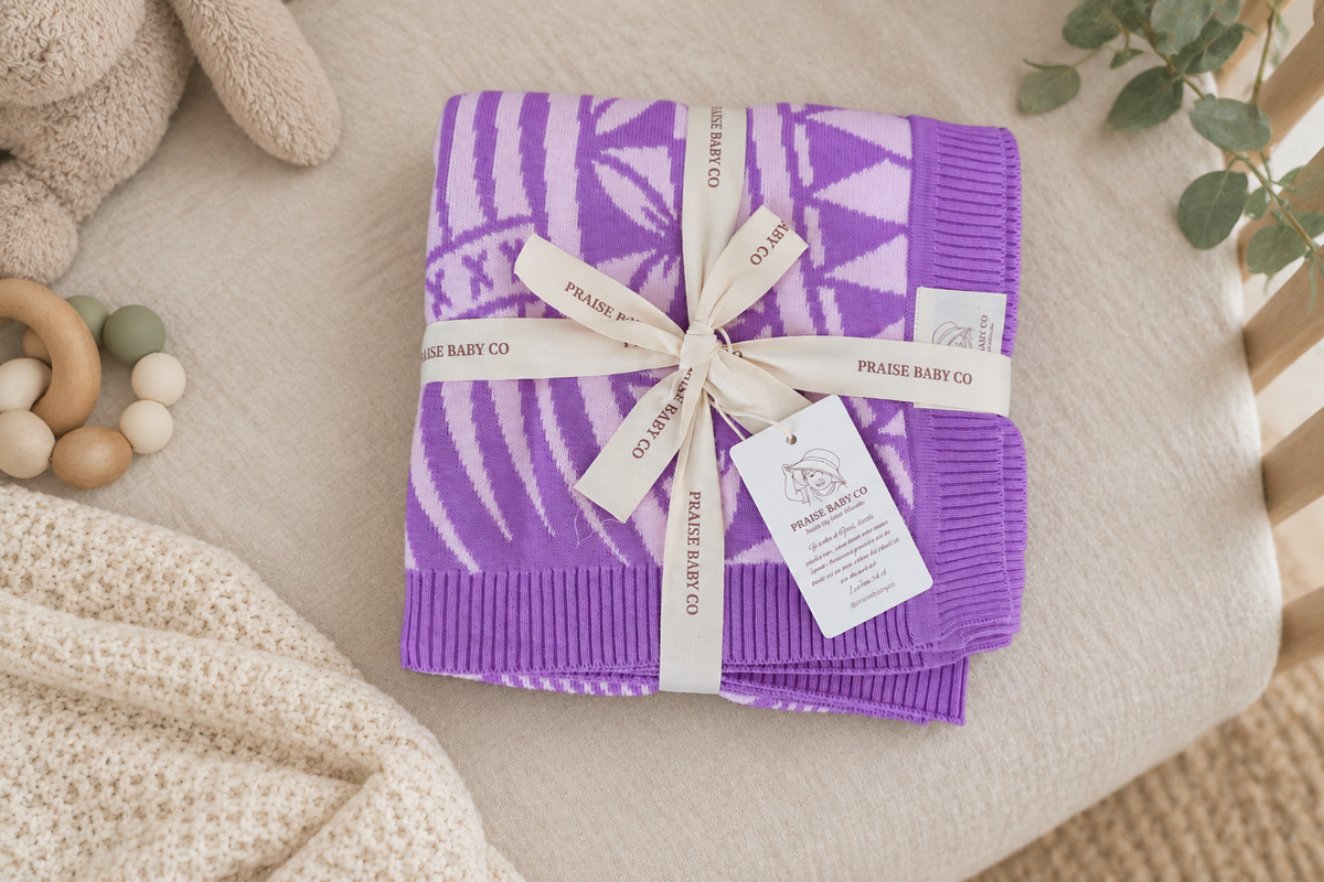 A folded purple and white patterned knit baby blanket tied with a cream ribbon and tag, styled on a beige fabric surface with soft nursery props.