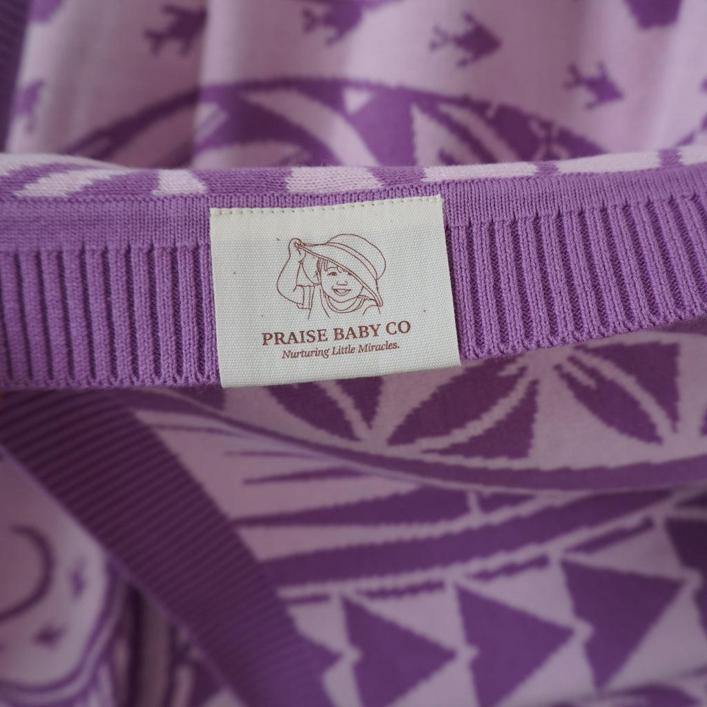A close-up of a purple and white patterned knit garment held by a hand, showing a white sewn-in label that reads 'PRAISE BABY CO' with a baby illustration.